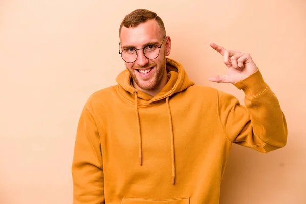 Young Caucasian Man Isolated Beige Background Holding Something Little Forefingers — Stock Photo, Image