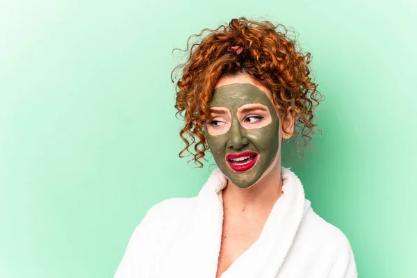 Young red hair woman with a facial aloe vera mask after a bath isolated looks aside smiling, cheerful and pleasant.