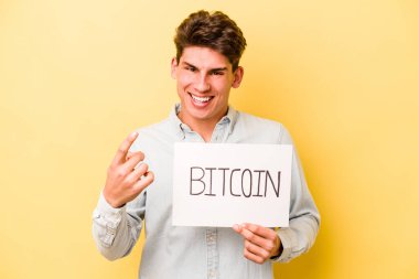 Elinde Bitcoin levhası olan genç beyaz bir adam sarı arka planda izole edilmiş seni işaret ediyor sanki davet ediyormuş gibi..