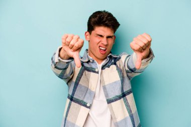 Mavi arka planda izole edilmiş genç beyaz bir adam baş parmağını aşağı gösteriyor ve hoşlanmadığını ifade ediyor..