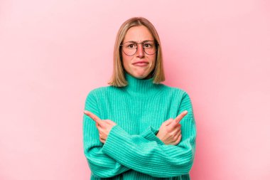 Pembe arka plan noktalarında izole edilmiş genç beyaz kadın iki seçenek arasında seçim yapmaya çalışıyor..