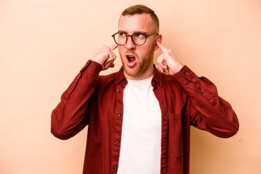 Beyazlı genç bir adam bej arka planda izole edilmiş parmaklarıyla kulaklarını kapatıyor, gürültülü bir ortam yüzünden stresli ve çaresiz..