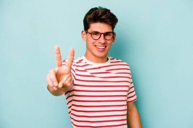 Mavi arka planda izole edilmiş genç beyaz adam zafer işareti gösteriyor ve gülümsüyor..