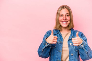 Pembe arka planda izole edilmiş genç beyaz kadın gülümsüyor ve başparmağını kaldırıyor.