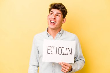 Sarı zemin üzerinde Bitcoin levhası tutan genç beyaz adam hedeflere ve amaçlara ulaşma hayali kuruyor.