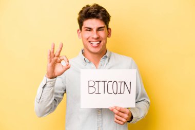 Elinde Bitcoin levhası olan beyaz bir genç adam sarı arka planda izole edilmiş neşeli ve kendinden emin bir şekilde iyi niyet gösteriyor..