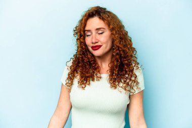 Mavi arka planda izole edilmiş genç beyaz kadın gülüyor ve gözlerini kapıyor, rahatlamış ve mutlu hissediyor..