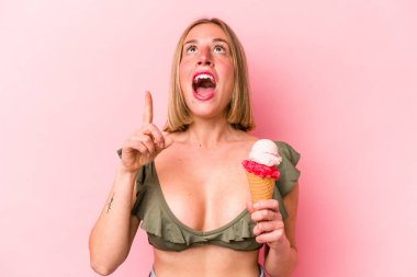 Genç beyaz kadın bikini giyiyor ve elinde pembe arka planda izole edilmiş ağzı açık bir dondurma tutuyor..