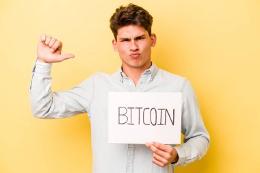 Sarı arka planda izole edilmiş bitcoin pankart tutan genç beyaz adam gururlu ve kendinden emin hissediyor, örnek olarak.