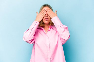 Mavi arka planda izole edilmiş genç beyaz kadın elleriyle gözlerini kapatmaktan korkuyor..