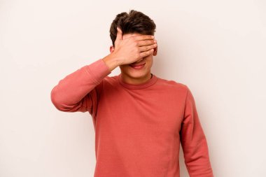 Beyaz arka planda soyutlanmış beyaz tenli genç bir adam elleriyle gözlerini kapatıyor, geniş bir gülümsemeyle bir sürpriz bekliyor..