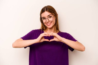Beyaz arka planda izole edilmiş genç beyaz kadın gülümsüyor ve elleriyle kalp şekli gösteriyor..
