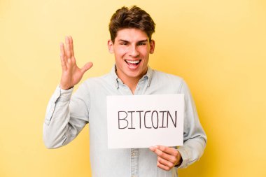 Sarı arka planda Bitcoin pankartı tutan genç beyaz adam hoş bir sürpriz alıyor, heyecanlı ve el kaldırıyor..