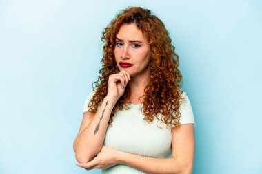 Kızıl saçlı genç bir kadın mavi arka planda izole edilmiş düşünüyor ve yukarı bakıyor, yansıtıyor, düşünüyor, hayal kuruyor..