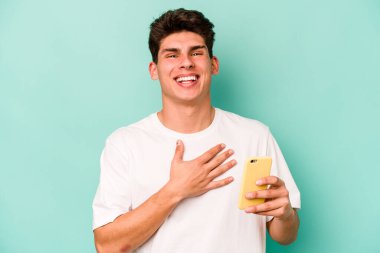 Mavi arka planda cep telefonu tutan genç beyaz adam elini göğsünde tutarak kahkahalar atıyor..