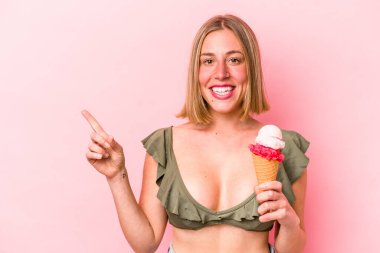 Bikini giyen ve pembe arka planda izole edilmiş dondurma tutan beyaz bir kadın gülümsüyor ve boş alanda bir şeyler gösteriyor..