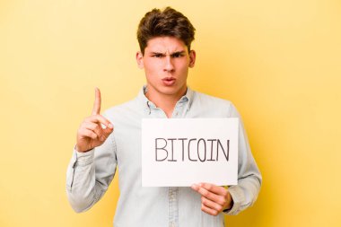Elinde bitcoin levhası olan genç beyaz bir adam sarı arka planda izole edilmiş harika bir fikre sahip, yaratıcılık kavramı..