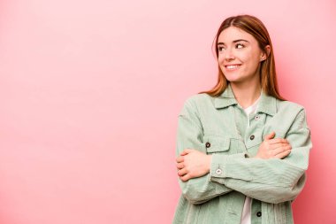 Pembe arka planda izole edilmiş genç beyaz kadın kollarını kavuşturarak gülümsüyor..