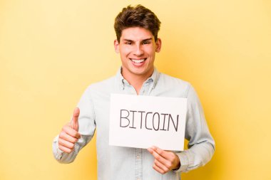 Sarı arka planda izole edilmiş bitcoin pankart tutan genç beyaz adam gülümsüyor ve başparmağını kaldırıyor.