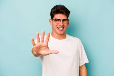 Mavi arka planda izole edilmiş genç beyaz adam gülümseyerek beş numarayı parmaklarıyla gösteriyor..
