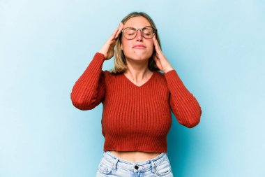 Mavi arka planda izole edilmiş genç beyaz kadın şakaklara dokunuyor ve baş ağrısı çekiyor..