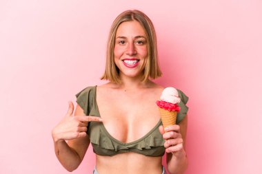 Bikini giyen beyaz bir kadın pembe arka planda izole edilmiş bir dondurma tutuyor. Elle bir gömlek fotokopi alanını işaret ediyor. Gururlu ve kendinden emin.