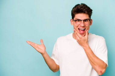 Mavi arka planda izole edilmiş genç beyaz bir adam avucunun içinde fotokopi alanı tutuyor, el üstünde tutuyor. Şaşırmış ve mutlu..