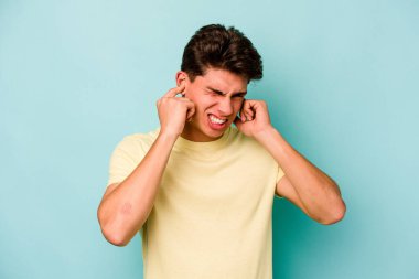 Mavi arka planda izole edilmiş genç beyaz adam kulaklarını elleriyle kapatıyor..