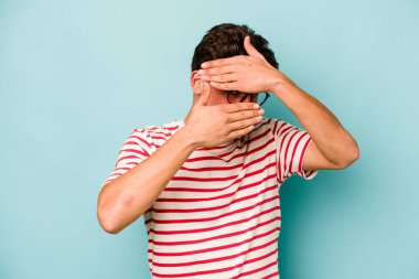 Mavi arka planda izole edilmiş genç beyaz adam kameraya parmaklarıyla göz kırpıyor, yüzünü kapatmaktan utanıyor..