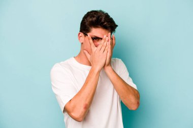 Mavi arka planda izole edilmiş genç beyaz adam kameraya parmaklarıyla göz kırpıyor, yüzünü kapatmaktan utanıyor..