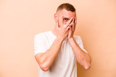 Beyazlı genç bir adam bej arka planda izole edilmiş. Kameraya parmaklarıyla göz kırpıyor, yüzünü kapatmaktan utanıyor..