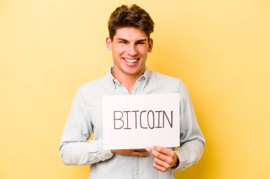 Sarı arka planda izole edilmiş bitcoin pankart tutan genç beyaz adam kahkahalar atıyor ve elini göğsüne koyuyor..