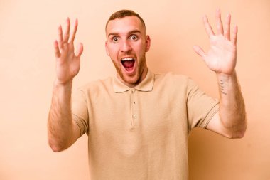 Beyazlı genç bir adam bej arka planda izole edilmiş bir şekilde zaferi veya başarıyı kutluyor. Şaşırmış ve şok olmuş..
