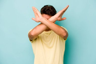 Kafkasyalı genç adam mavi arka planda izole edilmiş iki kolu çapraz tutuyor, inkar kavramı.