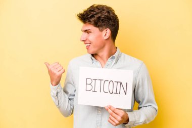 Elinde Bitcoin levhası olan genç beyaz bir adam sarı arka plan noktalarına izole edilmiş, başparmak uzakta, gülen ve kaygısız..