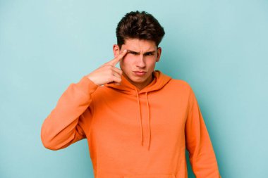 Mavi arka planda izole edilmiş genç beyaz bir adam şakağı parmağıyla işaret ediyor, düşünüyor ve bir göreve odaklanıyor..