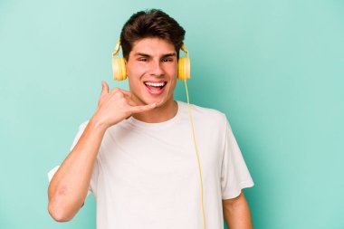 Mavi arka planda izole edilmiş müzik dinleyen genç beyaz adam parmaklarıyla bir cep telefonu çağrısı hareketi gösteriyor..