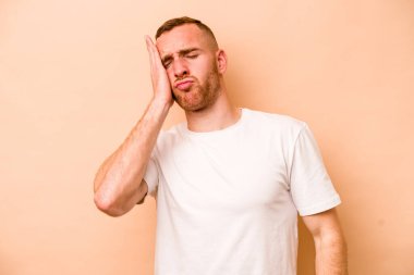 Beyazlı genç bir adam bej arka planda izole edilmiş. Yorgun ve uykulu. Elini başının üstünde tutuyor..