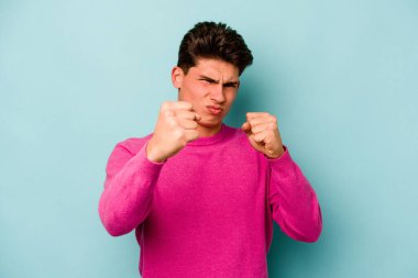 Mavi arka planda izole edilmiş genç beyaz adam yumruğunu kameraya gösteriyor, agresif yüz ifadesi var..
