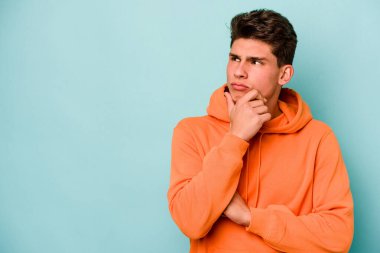 Mavi arka planda izole edilmiş genç beyaz adam şüpheli ve şüpheci bir ifadeyle yana bakıyor..