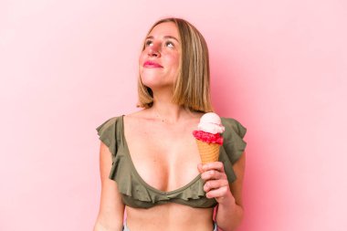 Bikini giyen beyaz bir kadın pembe arka planda izole edilmiş dondurma tutuyor. Amaçlarına ve amaçlarına ulaşma hayali kuruyor.
