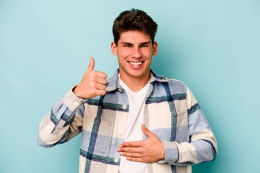 Mavi arka planda izole edilmiş genç beyaz adam karnına dokunuyor, nazikçe gülümsüyor, yemek yiyor ve tatmin oluyor..