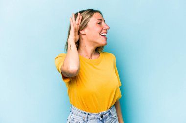 Mavi arka planda izole edilmiş genç beyaz kadın bir dedikoduyu dinlemeye çalışıyor..