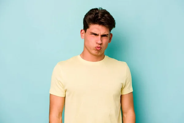Young caucasian man isolated on blue background blows cheeks, has tired expression. Facial expression concept.