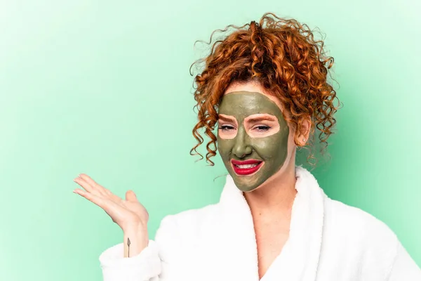 Young red hair woman with a facial aloe vera mask after a bath isolated showing a copy space on a palm and holding another hand on waist.