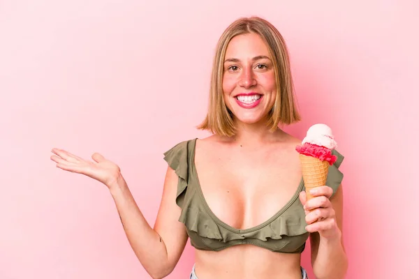 年轻的高加索女人穿着比基尼 拿着用粉色背景隔开的冰淇淋 手掌上有一个复制的空间 另一只手挽着腰部 — 图库照片