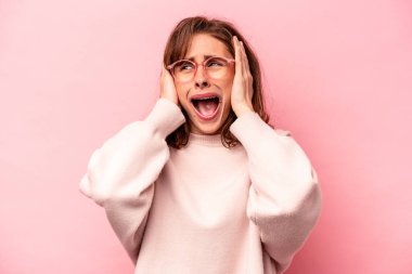 Pembe arka planda izole edilmiş genç, beyaz bir kadın. Kulakları kulaklarıyla kapatıyor. Çok fazla ses duymamaya çalışıyor..