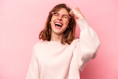 Pembe arka planda izole edilmiş genç beyaz kadın zaferi, tutkuyu ve coşkuyu kutluyor, mutlu bir ifade..