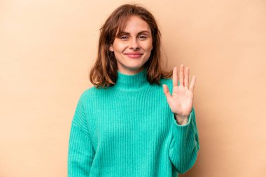 Beyaz tenli genç bir kadın bej arka planda izole edilmiş. Gülümsüyor. Beş numarayı parmaklarıyla gösteriyor..