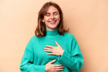 Beyaz tenli genç bir kadın bej arka planda izole edilmiş mutlu bir şekilde gülüyor ve ellerini karnının üstünde tutarak eğleniyor..